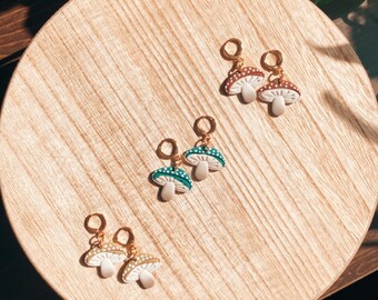 May include: Three pairs of mushroom-shaped earrings on a wooden surface. The earrings are gold-toned and feature white mushroom caps with brown, green, and white spots.