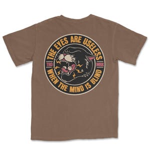 A traditional tattoo shirt featuring a panther with eyes in its mouth and the phrase “The eyes are useless when the mind is blind,” printed on a Comfort Colors tee for fans of tattoo art and alternative clothing.