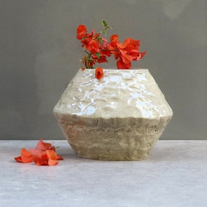 以下が含まれることがあります： テクスチャ加工された表面と幾何学的な形をした白いセラミックの花瓶。花瓶にはオレンジ色の花が活けてあります。