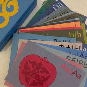 Könnte beinhalten: Eine blaue Schachtel mit goldfarbenen Buchstaben und ein Stapel bunter Alphabetkarten. Die Karten zeigen verschiedene Buchstaben und Illustrationen, darunter einen roten Apfel mit einem dekorativen Design. Der Text auf der Schachtel lautet "Qazaq Alphabet".
