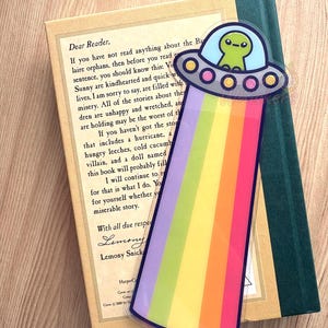 Puede incluir: Un marcapáginas colorido con un extraterrestre verde en un OVNI, con un rayo arcoíris. El marcapáginas está colocado sobre un libro con el texto "Dear Reader" y "Lemony Snicket".