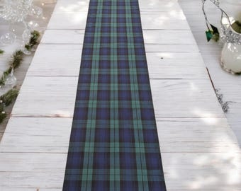 May include: A long, rectangular table runner featuring a classic plaid pattern in shades of deep navy blue, forest green, and thin black lines. The fabric is laid flat, showcasing a repeating geometric grid design across its entire visible length.