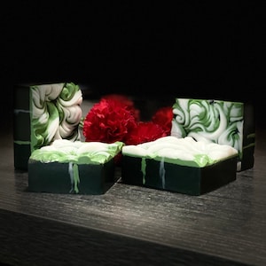 May include: Four bars of handmade soap with green and white swirls. The soap is on a dark wood surface with a red carnation flower.