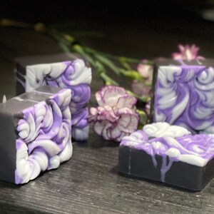 Pode incluir: Quatro barras de sabonete feitas &agrave; m&atilde;o com um design de redemoinho em tons de roxo e branco. As barras de sabonete est&atilde;o sobre uma superf&iacute;cie cinza escura.