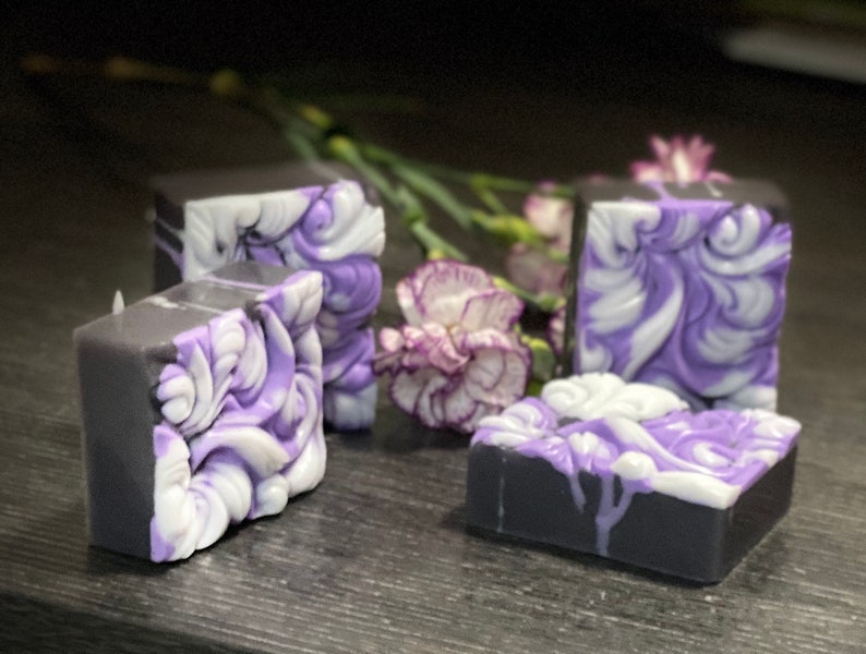 Pode incluir: Quatro barras de sabonete feitas &agrave; m&atilde;o com um design de redemoinho em tons de roxo e branco. As barras de sabonete est&atilde;o sobre uma superf&iacute;cie cinza escura.