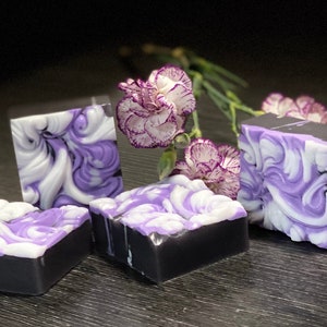 Pode incluir: Quatro barras de sabonete artesanal com um design de redemoinho em tons de roxo e branco. O sabonete est&aacute; colocado sobre uma superf&iacute;cie de madeira escura com flores roxas.