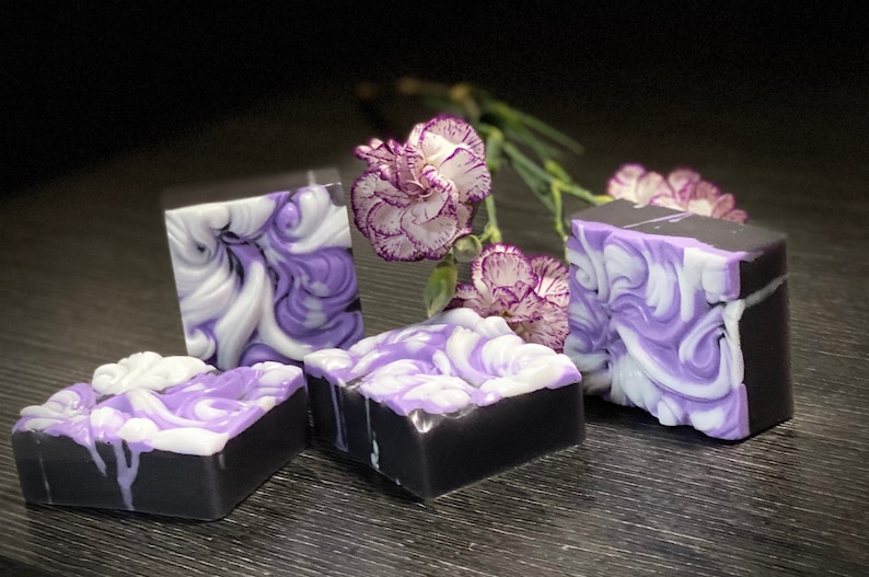 Pode incluir: Quatro barras de sabonete artesanal com um design de redemoinho em tons de roxo e branco. O sabonete est&aacute; colocado sobre uma superf&iacute;cie de madeira escura com flores roxas.