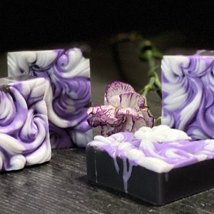 May include: Four handmade soap bars with a swirl design in shades of purple and white. The soap bars are on a dark gray surface with a single purple flower.