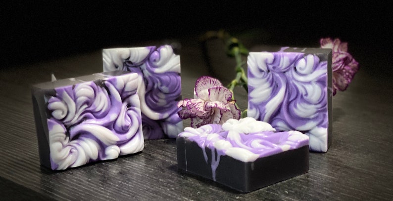 Pode incluir: Quatro barras de sab&atilde;o feitas &agrave; m&atilde;o com um design de redemoinho em tons de roxo e branco. As barras de sab&atilde;o est&atilde;o sobre uma superf&iacute;cie cinza escura com uma &uacute;nica flor roxa.
