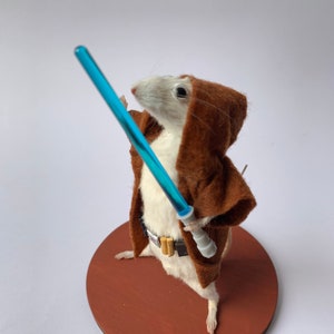 Peut inclure: Une souris naturalisée vêtue en Jedi, tenant un sabre laser bleu. La souris porte une robe à capuche marron et une ceinture avec des boucles dorées. Elle se tient sur une base ronde en bois brun rougeâtre sur fond blanc.