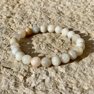 Puede incluir: Un brazalete de piedra lunar blanca con una mezcla de cuentas grises claras y oscuras. El brazalete está hecho de piedras naturales y tiene un diseño simple.