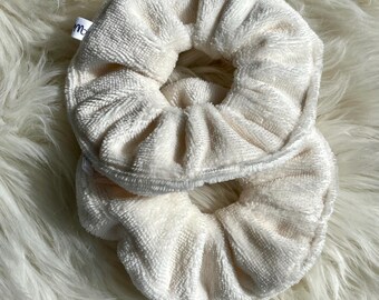 Puede incluir: Dos scrunchies de color blanco roto hechos de tela suave y peluda. Los scrunchies están apilados uno encima del otro.