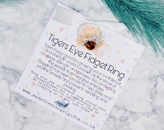 May include: A white card with pink and blue polka dots and the text "Tigers Eye Fidget Ring" in black. The card describes the benefits of wearing a tiger's eye ring. A silver ring with a brown tiger's eye stone is on top of the card.