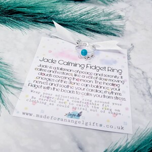May include: A silver and turquoise jade calming fidget ring with a white ribbon and a card that reads "Jade Calming Fidget Ring" and "Jade is a talisman of peace and serenity. It calms and restores, like a veil of slow moving clouds covering the moon. The healing energies of this stone can balance your nerves and soothe your cardiac rhythms. Fidget with the beads to calm you from stress. Wrap your adjustable ring around your finger for a comfortable fit. Be careful not to catch the ring on clothing as could spiral out of shape. www.madeforanangelgifts.co.uk"