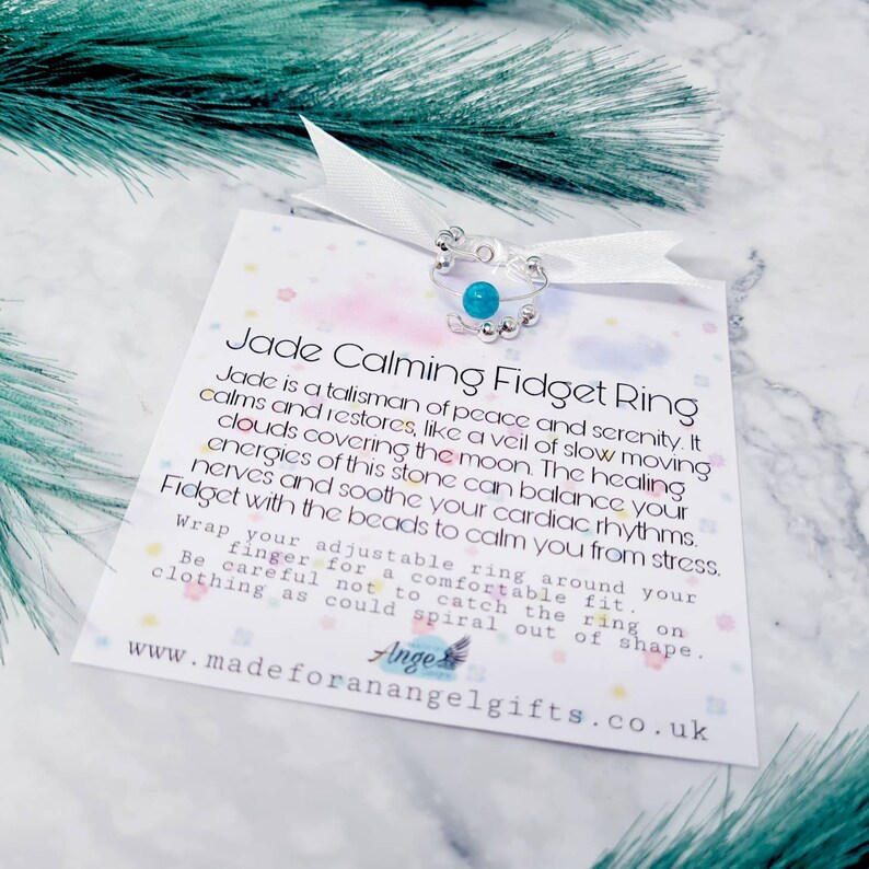 May include: A silver and turquoise jade calming fidget ring with a white ribbon and a card that reads "Jade Calming Fidget Ring" and "Jade is a talisman of peace and serenity. It calms and restores, like a veil of slow moving clouds covering the moon. The healing energies of this stone can balance your nerves and soothe your cardiac rhythms. Fidget with the beads to calm you from stress. Wrap your adjustable ring around your finger for a comfortable fit. Be careful not to catch the ring on clothing as could spiral out of shape. www.madeforanangelgifts.co.uk"