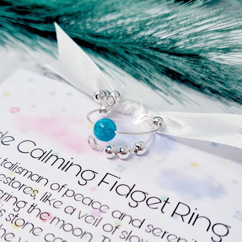 May include: A silver fidget ring with a turquoise bead and a small silver loop. The ring is on a white card with the text "Calming Fidget Ring" and a description of the ring.