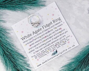May include: A silver fidget ring with white agate beads. The ring is on a white card with pink and green flowers. The card reads "White Agate Fidget Ring" and includes information about the properties of white agate.