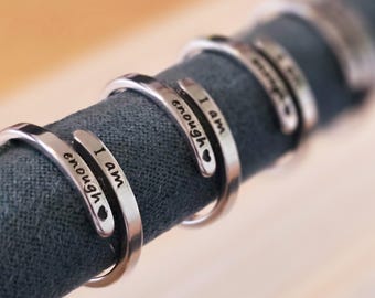 May include: Close-up of four silver rings with the words "I am enough" engraved on each. The rings are displayed on a gray fabric background.