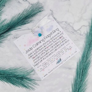 May include: A white card with the text "Jade Calming Fidget Ring" and a description of the benefits of jade. The card is next to a silver ring with a turquoise bead.