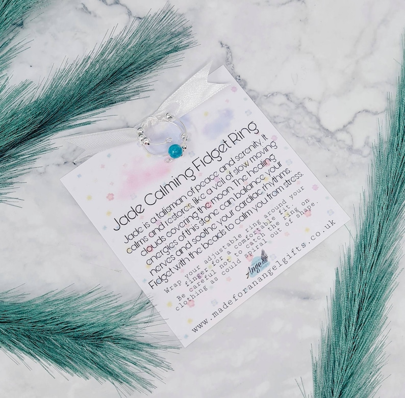 May include: A white card with the text "Jade Calming Fidget Ring" and a description of the benefits of jade. The card is next to a silver ring with a turquoise bead.