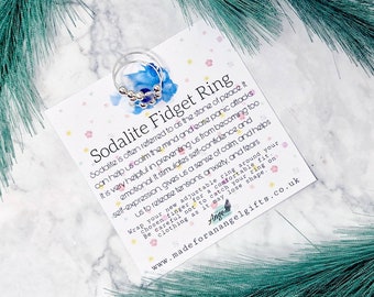 May include: A silver ring with blue sodalite beads. The ring is on a white card with the text "Sodalite Fidget Ring" and information about the benefits of sodalite.
