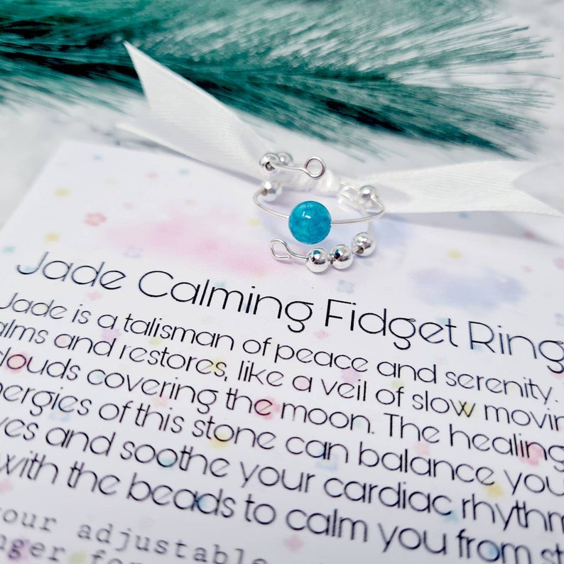 May include: A silver fidget ring with a turquoise jade stone and silver beads. The ring is on a white card with the text "Jade Calming Fidget Ring" and a description of the benefits of jade.