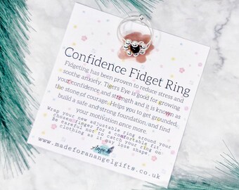 May include: Silver fidget ring with a brown tiger's eye stone. The card reads "Confidence Fidget Ring" and explains that fidgeting can reduce stress and anxiety. The card also mentions that tiger's eye is known for growing confidence and strength. The card is on a green background.