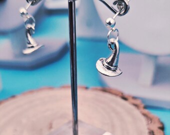 May include: Silver-toned witch hat earrings on display. The earrings are suspended from a silver post and feature a small, detailed witch hat charm. The display stand is clear, and the background is a blurred mix of blue and white. The text "Made for an Ange Designs" is visible.