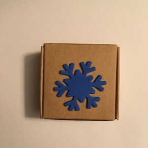 May include: A small, square, brown cardboard box with a blue snowflake shape affixed to the front. The snowflake is a solid, flat design. The box is closed and sits on a white surface.