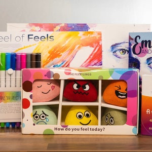 Może przedstawiać: Zestaw narzędzi edukacyjnych do nauki emocji. Zawiera wykres "Wheel of Feels", zestaw kolorowych markerów, pudełko pluszowych piłeczek emocji, książkę "Emotions and You" oraz zestaw kart "I've Got a Feeling..."
