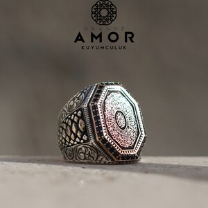 Op de afbeelding: Een zilveren ring met een achthoekig oppervlak met gedetailleerde kalligrafie en zwarte edelstenen. De ring heeft een gedetailleerd, gestructureerd ontwerp op de band en de tekst "GRANDE AMOR KUYUMCULUK" is zichtbaar.