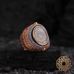 May include: A rose gold ring with an ornate design. The ring features a central blue stone with gold calligraphy and is surrounded by small blue gemstones. The ring is set on a dark, textured surface.