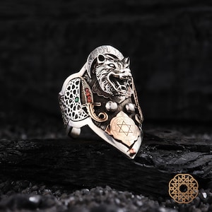 May include: A silver ring with a detailed wolf head design. The ring features gold accents, a Star of David emblem, and small red and green gemstones. The ring has an intricate, ornate design.