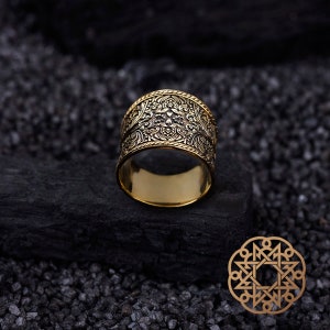 Puede incluir: Un anillo dorado con un intrincado diseño floral. El anillo ancho presenta un diseño detallado alrededor de la banda. Se muestra sobre una superficie oscura y texturizada, con un emblema decorativo en la esquina inferior derecha.
