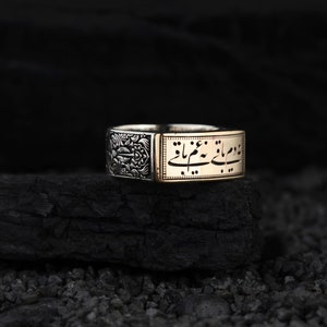 May include: A two-tone ring with detailed patterns. One section has a floral design, the other displays elegant script. The ring is set against a dark background, highlighting its design. The ring appears to be made of silver and gold.