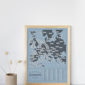 Puede incluir: Una impresión enmarcada de un mapa de Europa en tonos azules y grises, con códigos y nombres de ciudades. La impresión está en un marco de madera clara. El texto "The Great Places of Europe" está en la parte inferior.