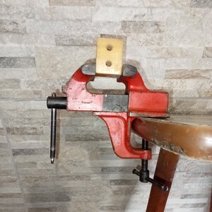 Puede incluir: Un tornillo de banco de metal rojo con una base de madera y un mango negro. El tornillo de banco está sujeto a una mesa de madera.