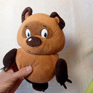 Peut inclure: Un ours en peluche marron avec des oreilles noires, un nez marron et des yeux blancs. L'ours est assis sur une surface blanche.