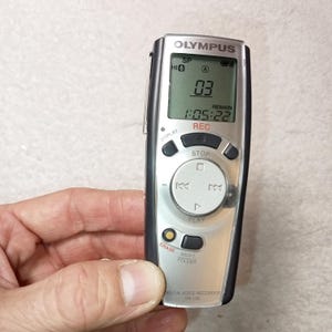 Może przedstawiać: Srebrny i czarny cyfrowy rejestrator głosu Olympus VN-120. Ekran LCD wyświetla "03" i "1:05:22". Funkcje obejmują przyciski nagrywania, zatrzymywania, odtwarzania, kasowania i menu. Z boku przymocowany jest srebrny klips.