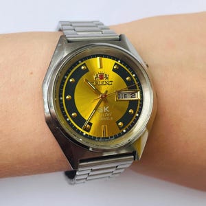 May include: A vintage Orient SK Crystal watch with a gold dial and a stainless steel band. The watch face has black accents and a day/date display. The watch is on a wrist.