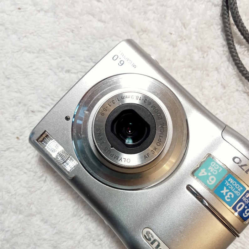 May include: Silver Olympus digital camera with a 6.0-megapixel resolution and 3x optical zoom. The camera features a lens with a 6.3-18.9mm zoom range and a built-in flash. The LCD screen is 6.4 cm.