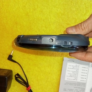 May include: A black and silver portable CD player with a headphone jack and a power button. The player is sitting on a yellow surface.