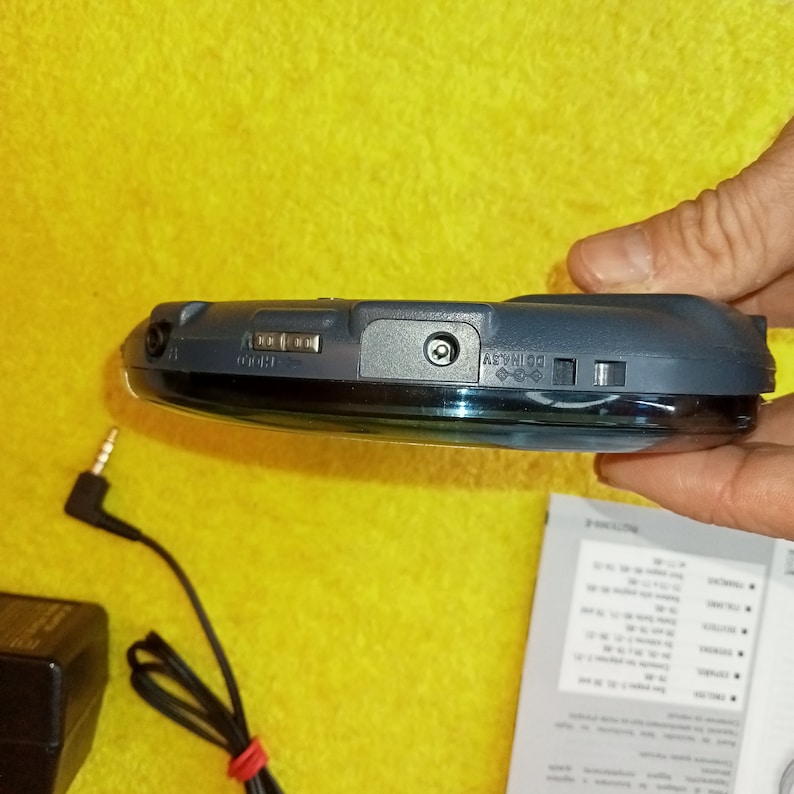 May include: A black and silver portable CD player with a headphone jack and a power button. The player is sitting on a yellow surface.