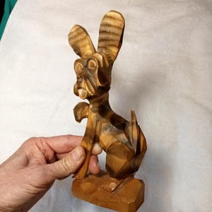 May include: A carved wooden dog figurine with large ears and a whimsical expression. The dog is brown with darker brown accents, and the carving style gives it a geometric appearance. The figurine is sitting on a small wooden base.
