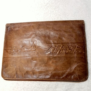 May include: A brown leather folio with an embossed airplane design and the text "AH-32". The folio has a rectangular shape and a slightly distressed appearance, suggesting it is vintage or has a rustic aesthetic.
