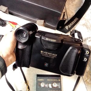 May include: A black Sharp Viewcam liquid crystal video camera with a black carrying case and a Panasonic strap. The camera has a lens, buttons, and ports. A VHS tape is also visible.