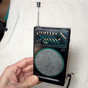 May include: A black Sony portable radio with a silver speaker cover and a green border. The radio has a retractable antenna and a black wrist strap. The radio's face has green and white markings. The word "SONY" is printed on the top right.