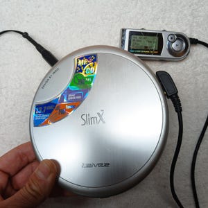 May include: A silver iRiver SlimX portable CD player with a circular design, featuring a holographic sticker with text including MP3, CD, and 55 hrs. A small silver remote control with a digital display is connected by a black cord to the main unit.