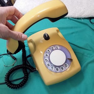 May include: A vintage yellow rotary dial telephone with a black coiled cord and a matching handset. The phone features a circular dial with numbers and a purple and white center. The handset is held up.