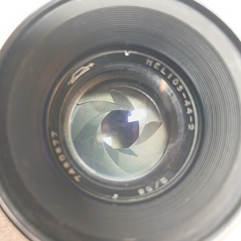 Peut inclure: Gros plan sur un objectif d'appareil photo avec le texte "HELIOS-44-2" et le num&eacute;ro "7460877" visibles. L'objectif a un ext&eacute;rieur noir et une ouverture avec plusieurs lamelles. Les lamelles d'ouverture sont partiellement ouvertes.